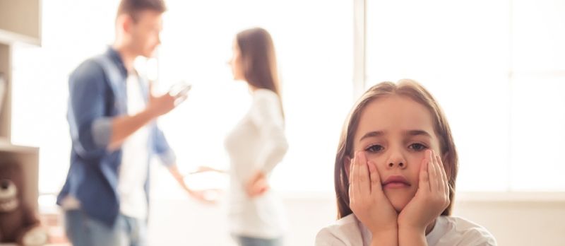 Trist liten jente ser på kameraet mens foreldrene hennes krangler i bakgrunnen