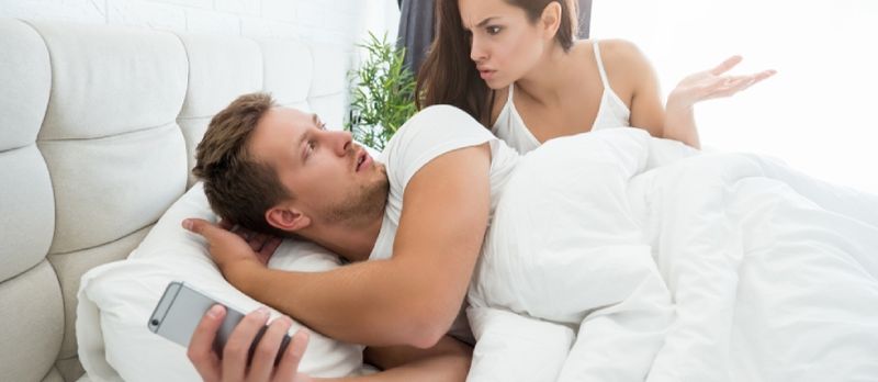 携帯電話を持つ若いカップルのハンサムな男とベッドに横たわっている間喧嘩している若い動揺した女性