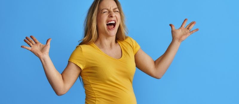 Mlada trudnica koja čeka bebu na izoliranoj plavoj pozadini, u ljutnji, ludo vrišti agresivnim izrazom lica i podignutim rukama