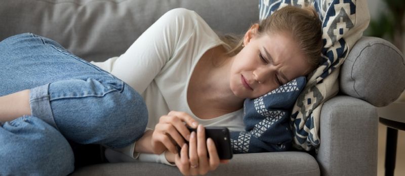 Narahat gənc qadın divanda uzanıb ağlayır, telefon tutaraq zəng mesajını gözləyir və smartfona baxır.