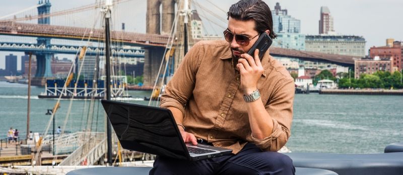 Radni čovjek za praćenje koji razgovara preko telefona i koristi prijenosno računalo Brooklyn Bridge u pozadini