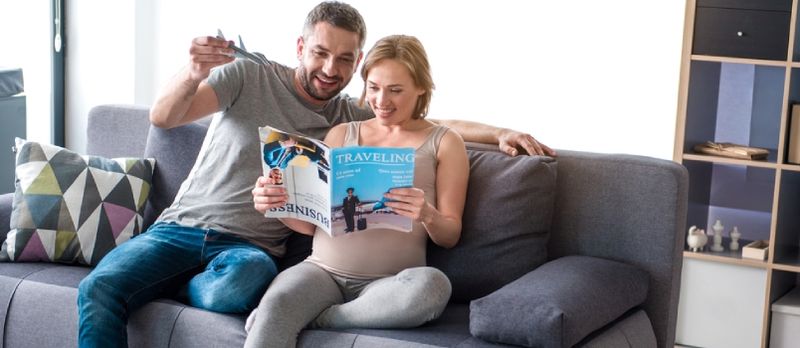 Planificarea călătoriei viitoare. Portret pe lungime întreagă al unui cuplu iubitor vesel care citește revista despre călătoria în timp ce stă pe canapea acasă