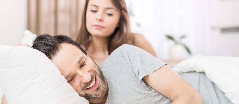 Eng jonk Koppel, déi am Bett läit. Ee vun hinnen benotzt den Telefon a flirtéiert