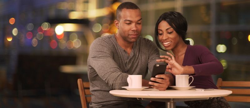 Cuplu negru fericit folosind telefonul inteligent în cafenea noaptea