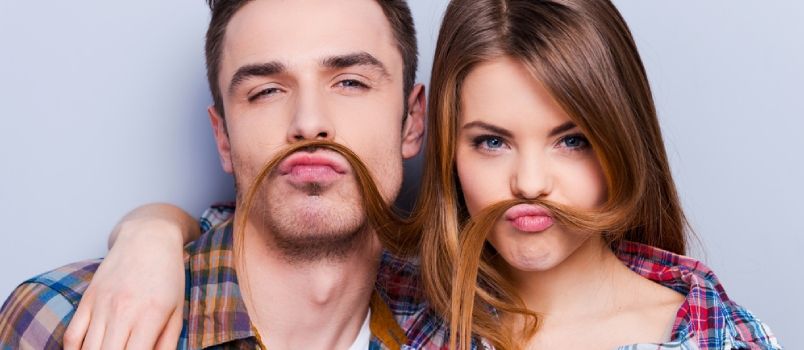 Càraid gaoil òg eireachdail a ’dèanamh mustache falsa bho fhalt fhad‘ s a tha iad nan seasamh an aghaidh cùl-fhiosrachadh liath
